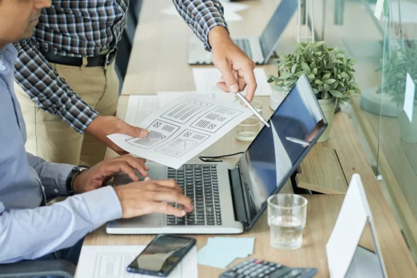 Two small business owners collaboratively reviewing website wireframes and mockups on paper, with one typing on a laptop, symbolizing the planning and development process for a new website. This image highlights the practical support offered in a free website starter kit for small businesses.