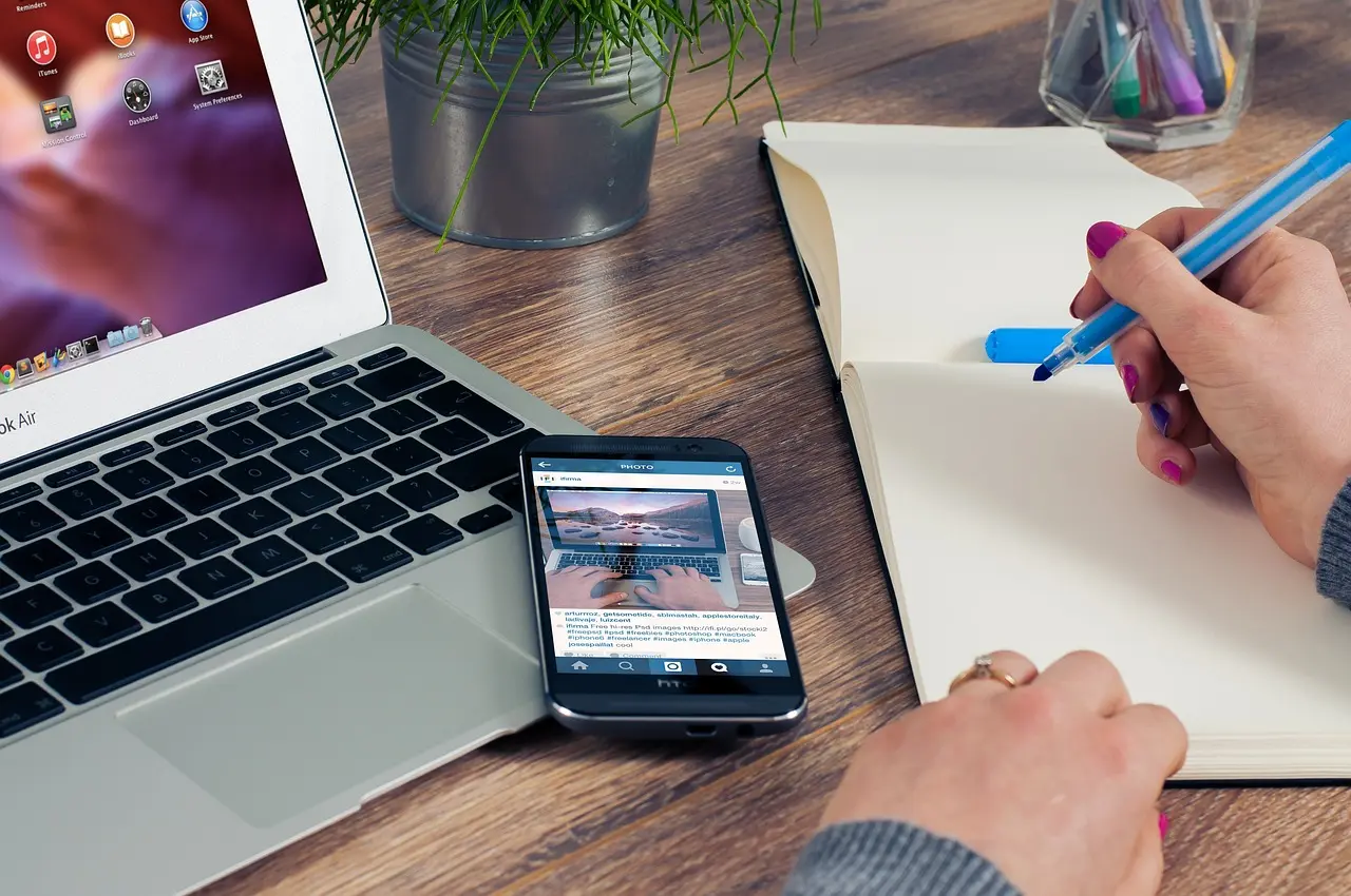 A person's hands writing in a notebook next to a laptop and smartphone displaying a website, symbolizing active learning and practical application from a free website guide download. This image represents the actionable insights provided by Launchpad Web Designs for building an effective online presence.