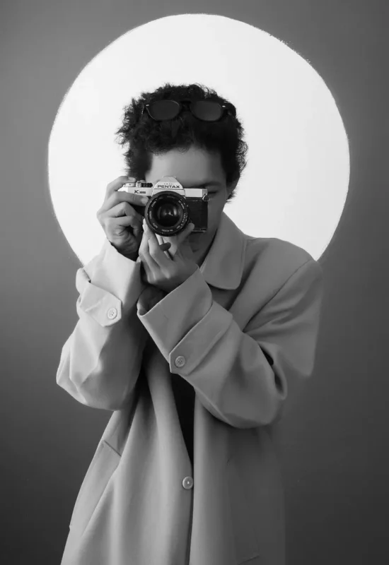 A professional photographer, viewed in a black and white portrait, holds a vintage Pentax camera to their face, ready to take a picture. A soft white light frames their head and shoulders against a dark background. This image represents a custom freelancer website design service tailored specifically for photographers, emphasizing a professional, artistic, and unique online portfolio.