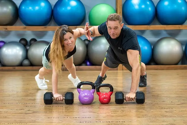 A personal trainer and his client, both in a plank position, are high-fiving. They are in a gym with exercise balls in the background and weights on the floor in front of them. This image is relevant for a website section on freelancer website design for personal trainers, virtual assistants, event planners, and translators.