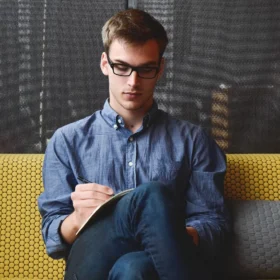 A focused freelancer, wearing glasses and a blue button-down shirt, sits on a modern yellow and gray couch, writing in a notebook. This image is ideal for illustrating articles about freelancer website designs and how to effectively showcase your professional skills online.