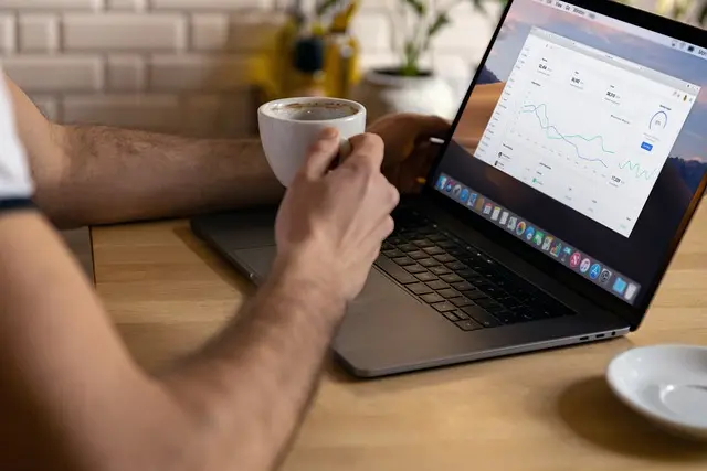 A person's hands hold a white coffee mug while they work on a laptop displaying a website analytics dashboard with line graphs and various data points. The screen shows a modern, clean interface for tracking website performance and traffic.