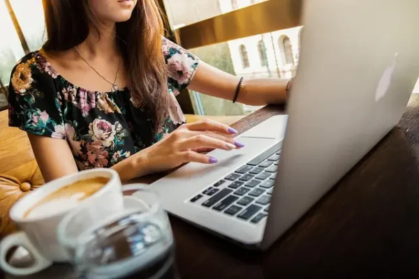 A female freelancer works remotely on her laptop in a sunlit cafe, with a coffee cup on the table next to her. The image represents the independent and flexible lifestyle and promotes the strategic benefits of Freelancer SEO as a vital tool for their online success.